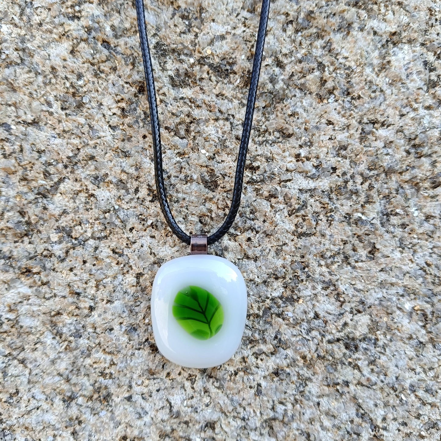 : A white glass pendant with a green leaf design hangs from a black cord necklace.