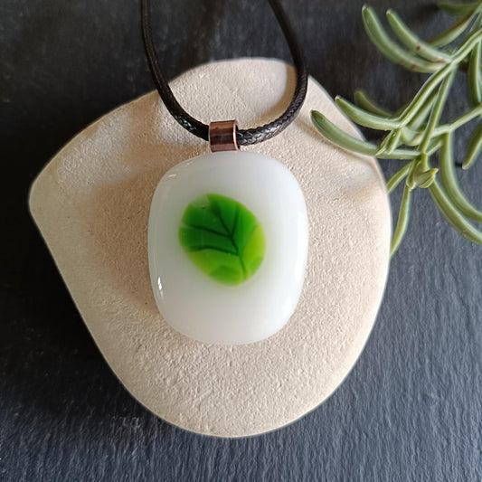 A white glass pendant with a green leaf design. The pendant is on a black cord and is resting on a light brown stone.