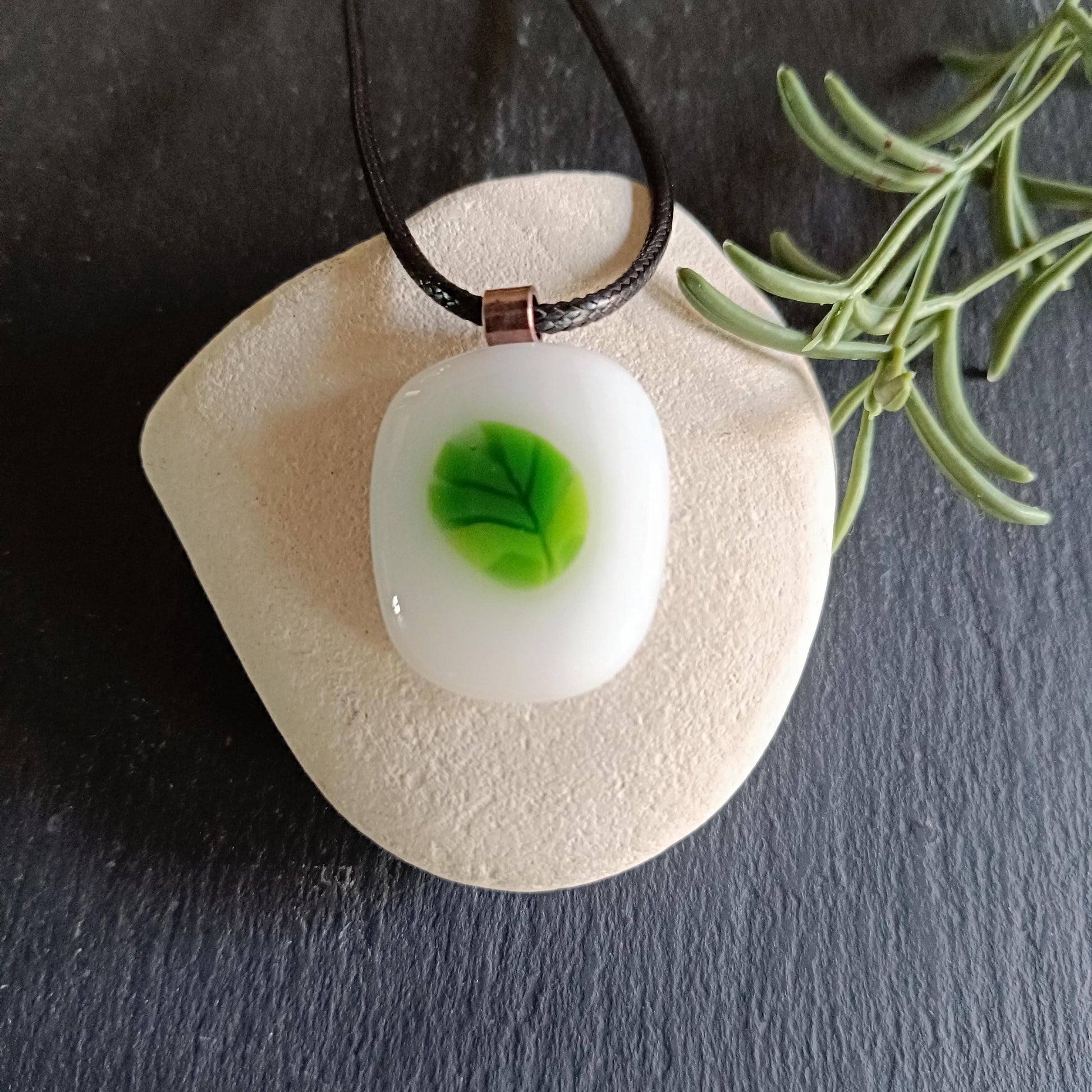 A white glass pendant with a green leaf design. The pendant is hanging from a black cord and is resting on a light brown stone.