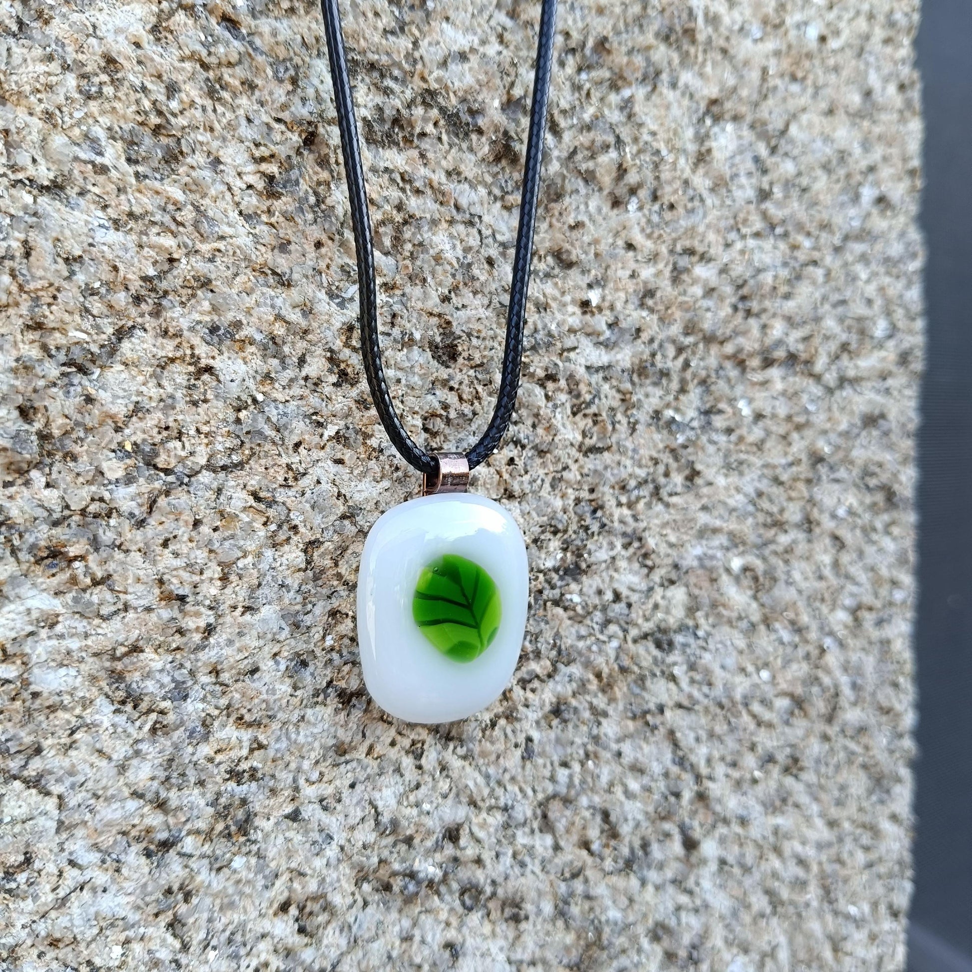 A white glass pendant with a green leaf design on a black cord necklace.