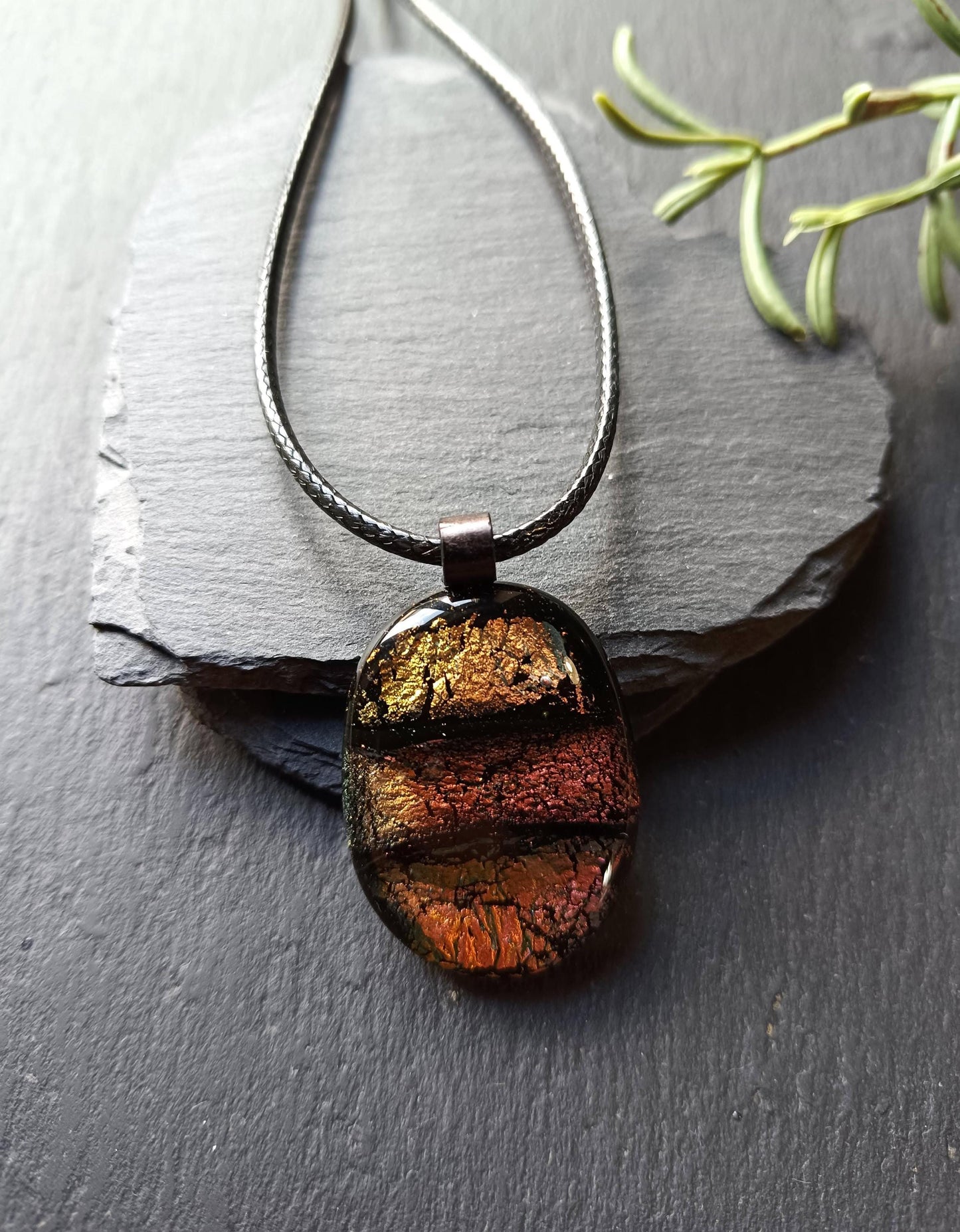 Oval handmade dichroic fused glass pendant in warm amber-copper tones on a black cord, layered design with metallic edges.