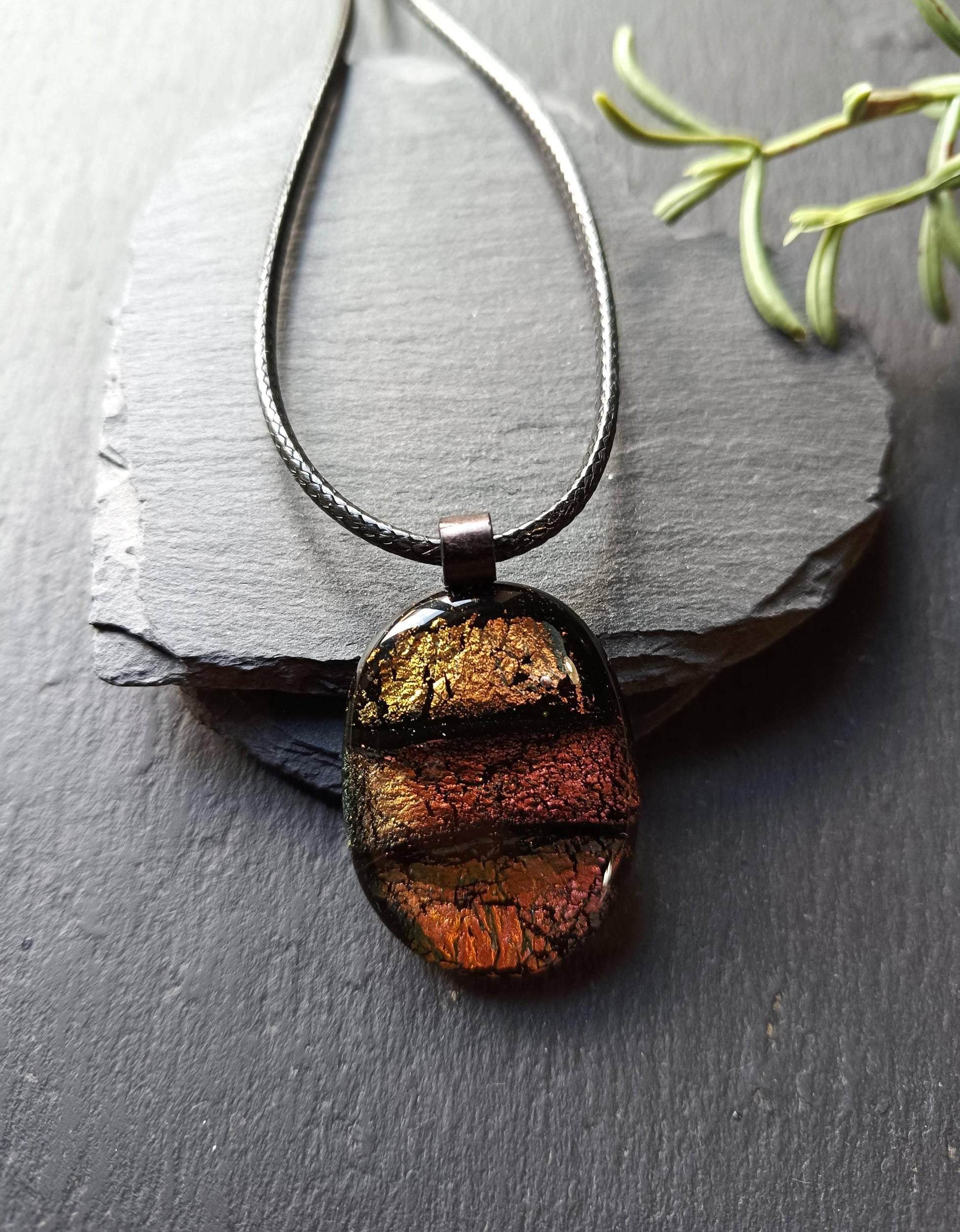 Oval handmade dichroic fused glass pendant in warm amber-copper tones on a black cord, layered design with metallic edges.