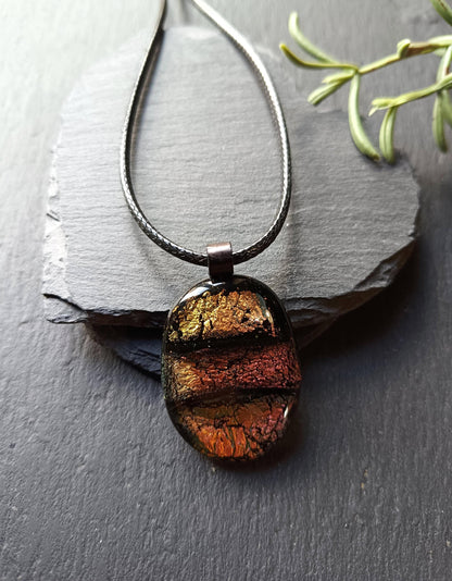 Oval handmade dichroic fused glass pendant in warm amber-copper tones on a black cord, layered design with metallic edges.