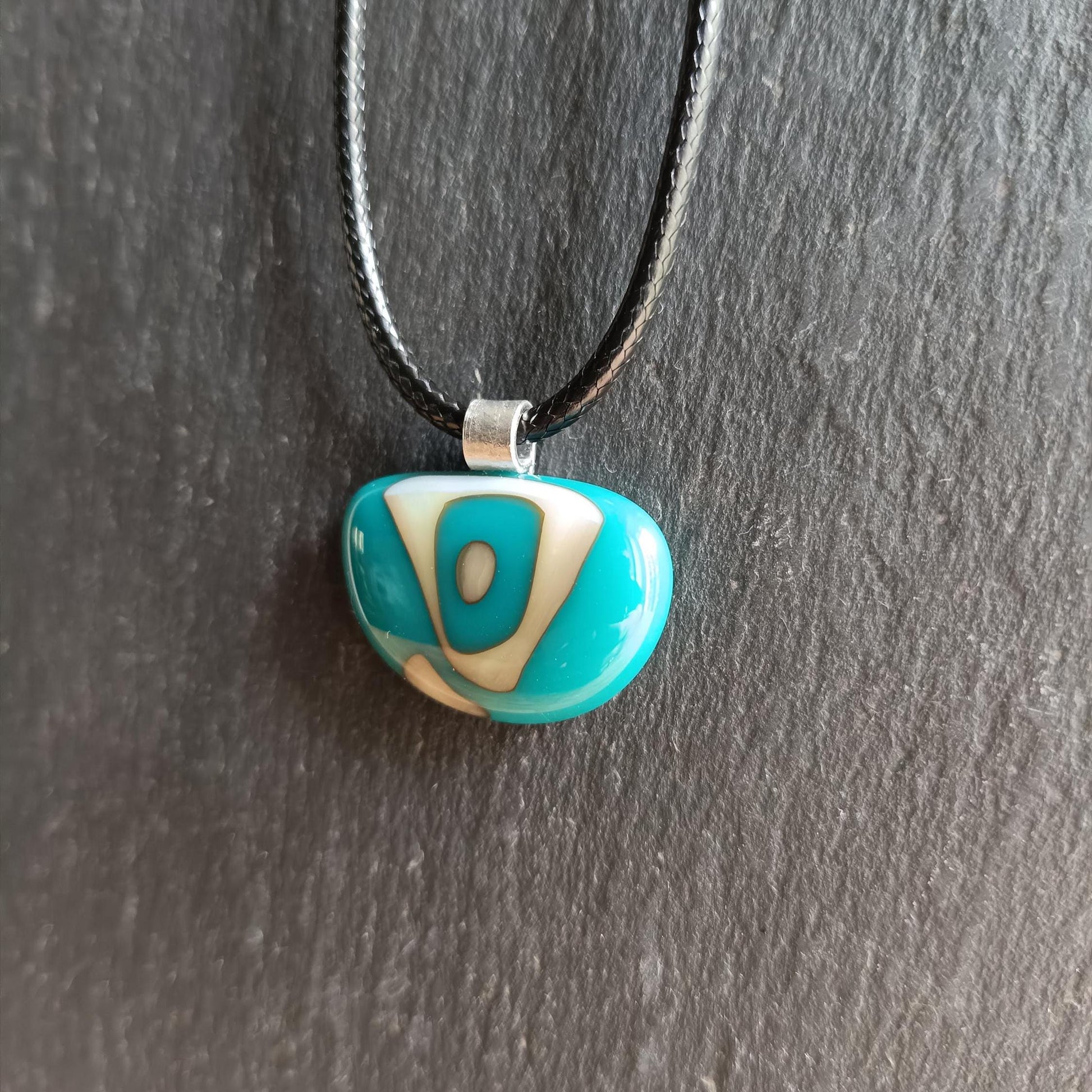 A turquoise and white glass pendant necklace with a black cord. The pendant is shaped semi circle and has a white abstract design.