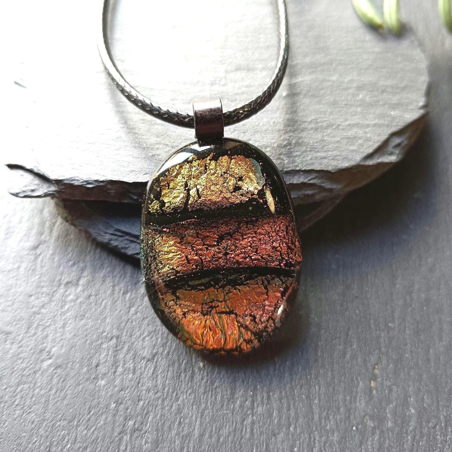 Oval handmade dichroic fused glass pendant in warm amber-copper tones on a black cord, layered design with metallic edges.