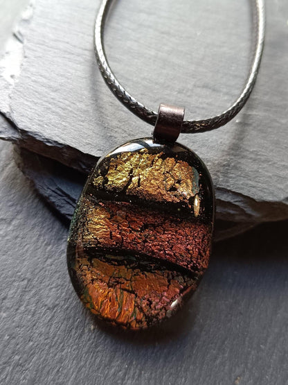 Oval handmade dichroic fused glass pendant in warm amber-copper tones on a black cord, layered design with metallic edges.