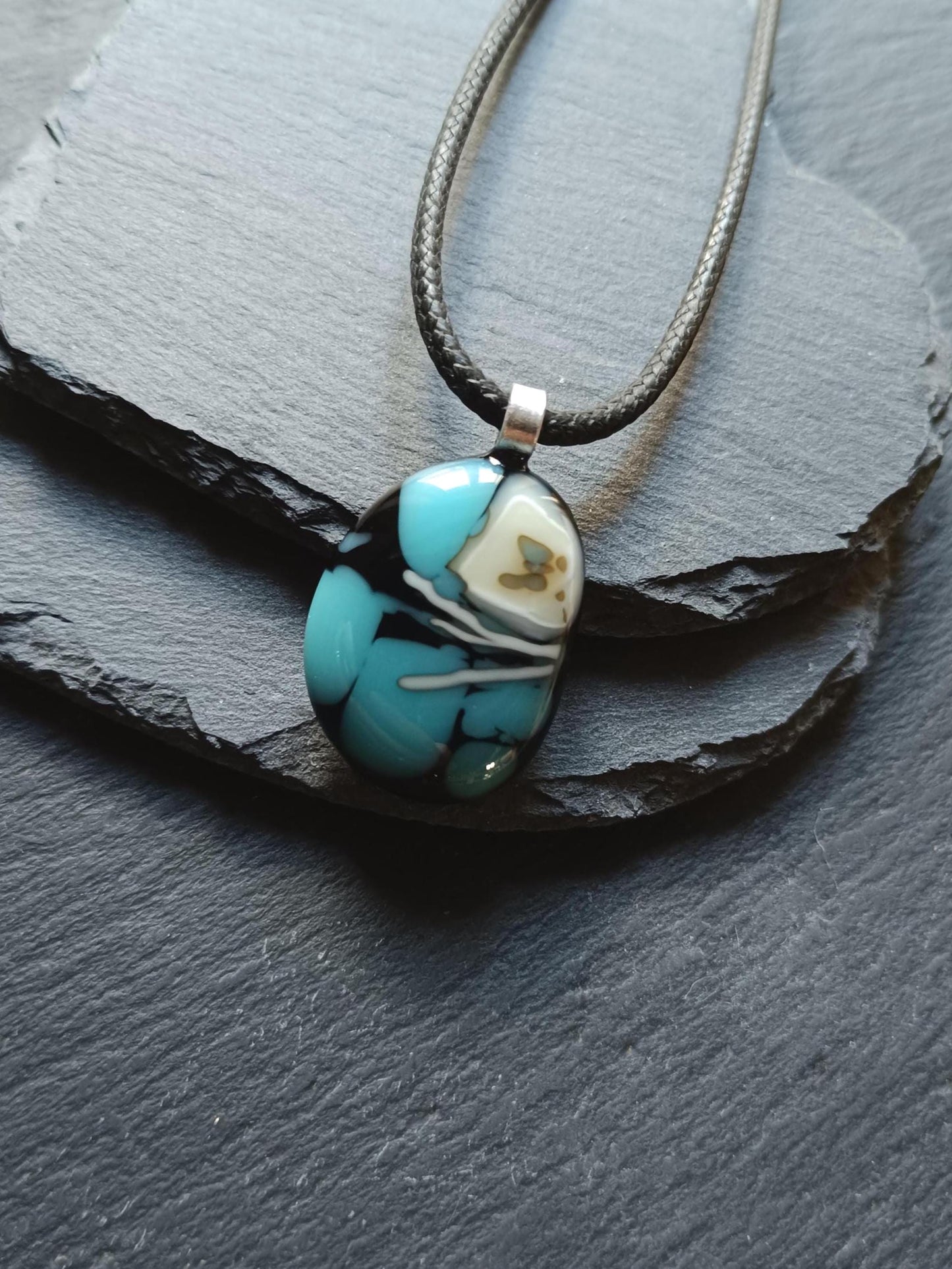 Fused glass pendant necklace in blue and black tones with white swirls and a silver bail on a black cord.