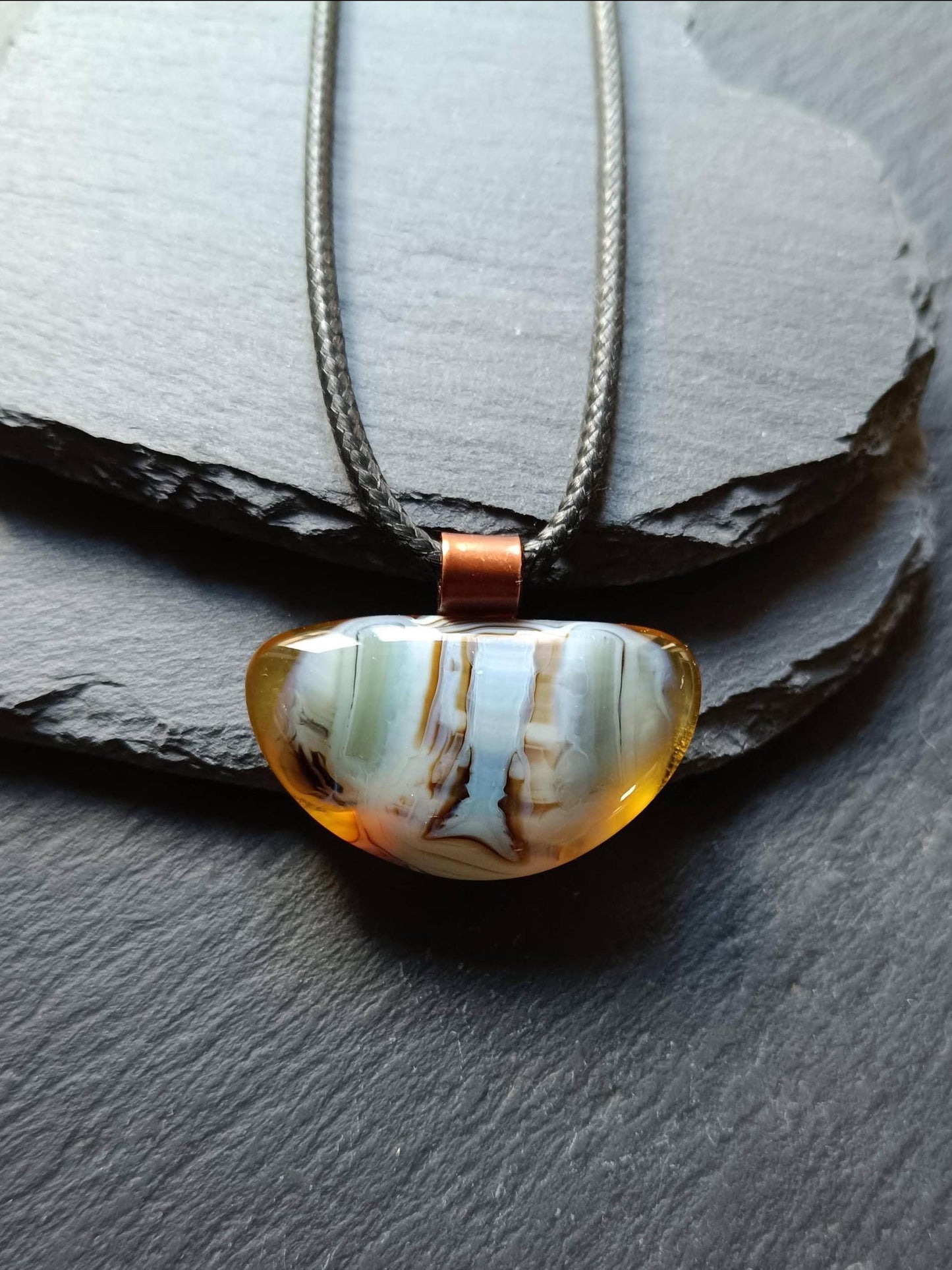Fused glass pendant on a grunge black cord necklace with copper bail; orange bottom with blue-green swirls and brown lines.
