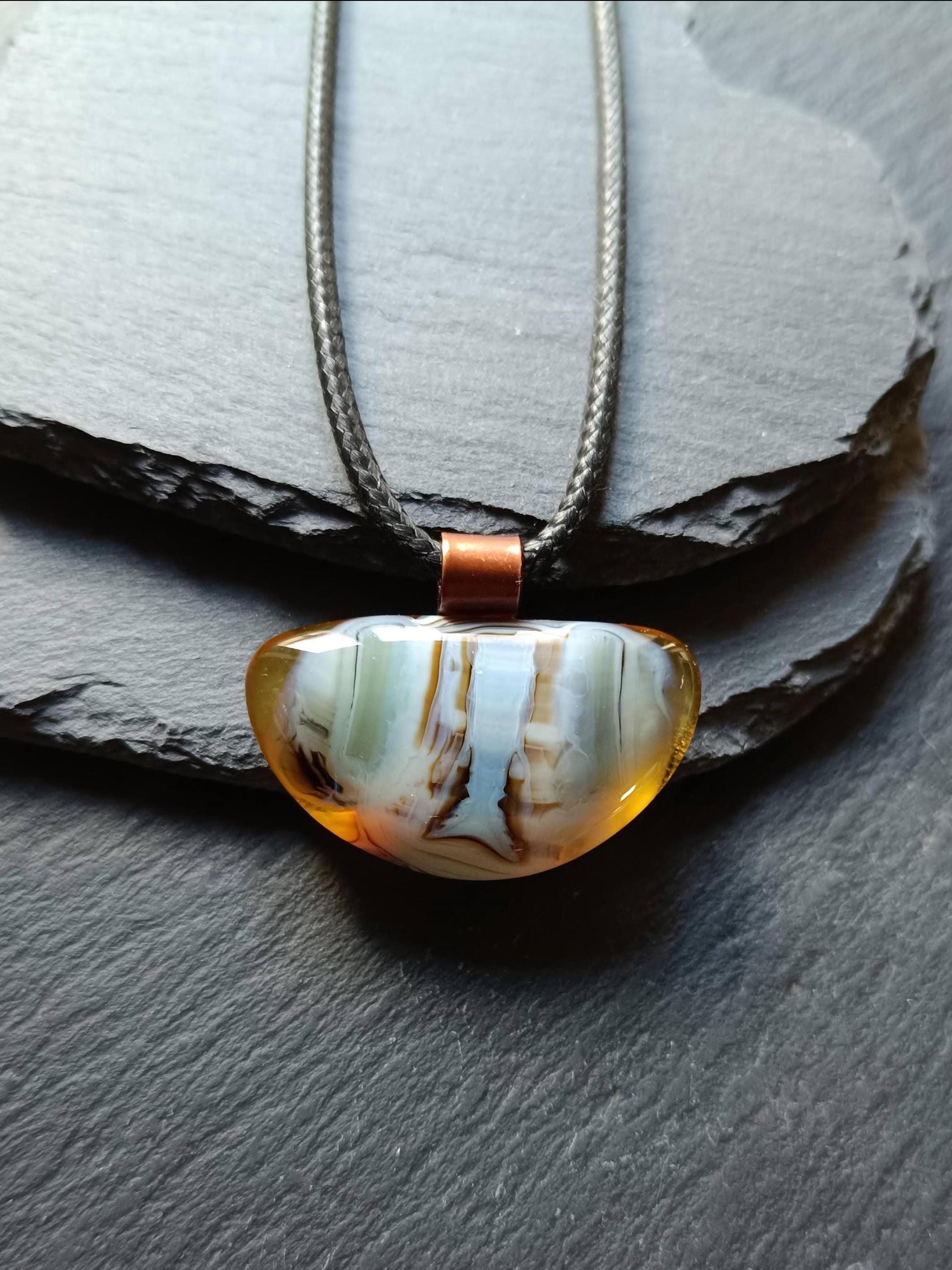 Fused glass pendant on a grunge black cord necklace with copper bail; orange bottom with blue-green swirls and brown lines.
