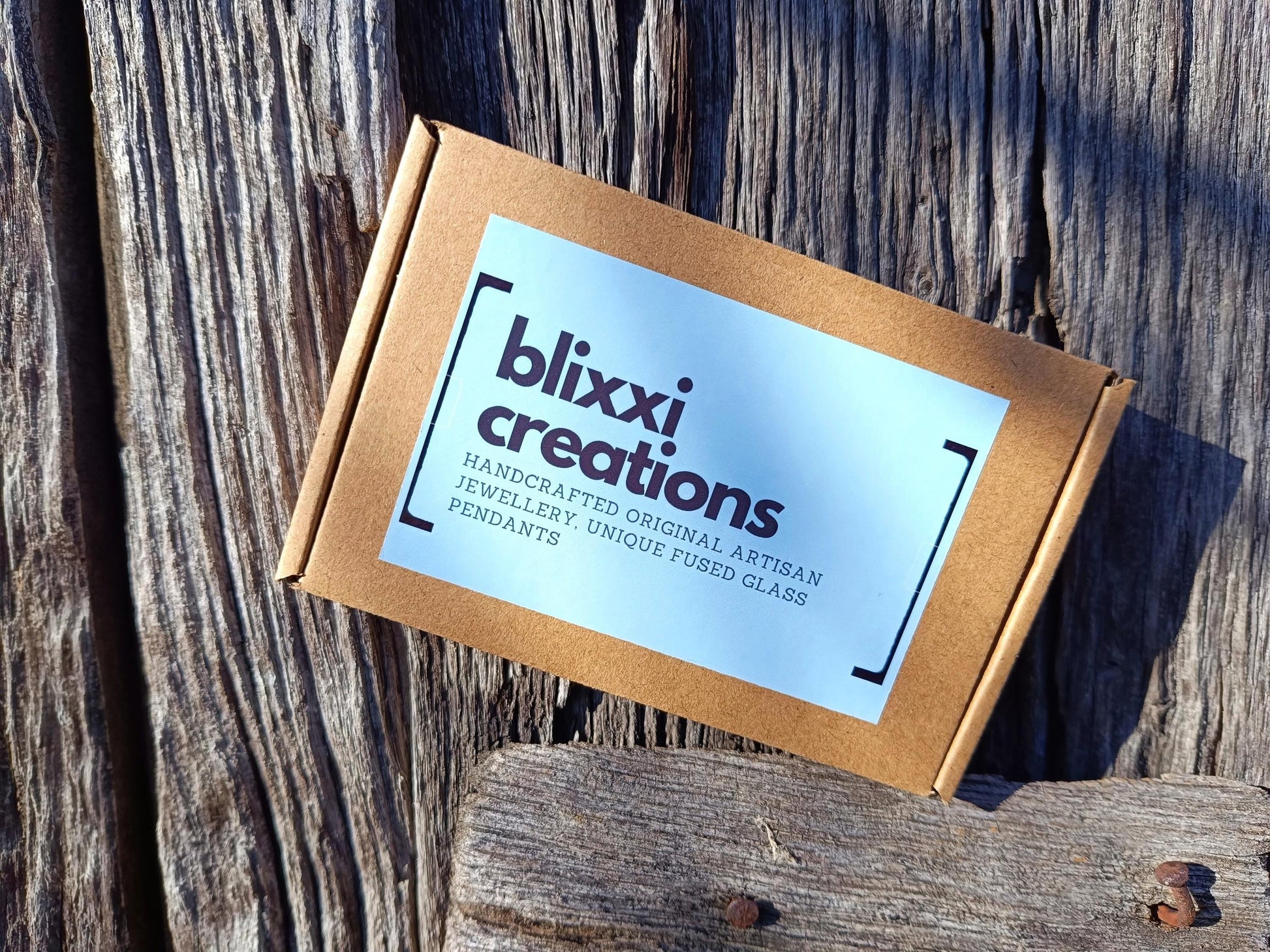 Fused glass orange pendant necklace packaging in a kraft box with blue label on rustic wood—handmade jewelry gift.