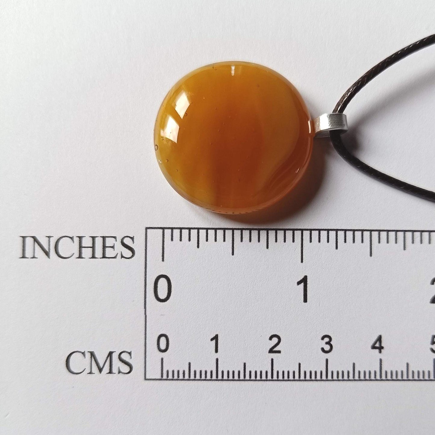 light warm coloured transparent glass pendant streaky on a cord necklace presented next to a ruler
