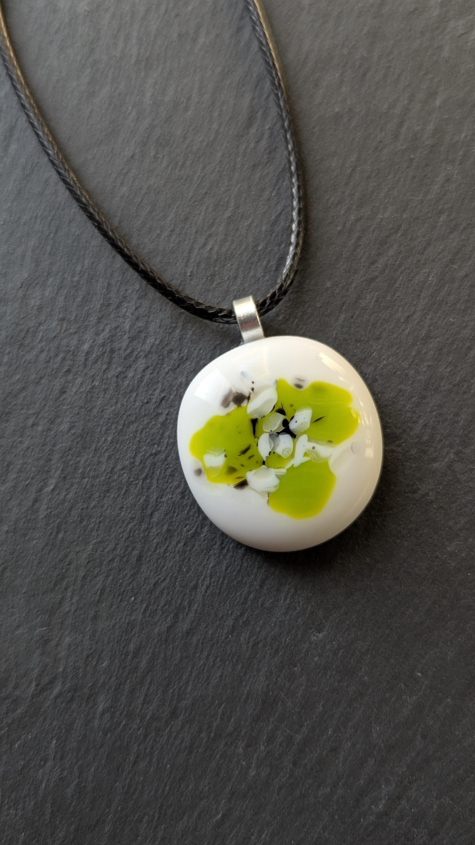 Round white fused-glass pendant with lime-green floral pattern on a silver bail, hanging from a black cord necklace.