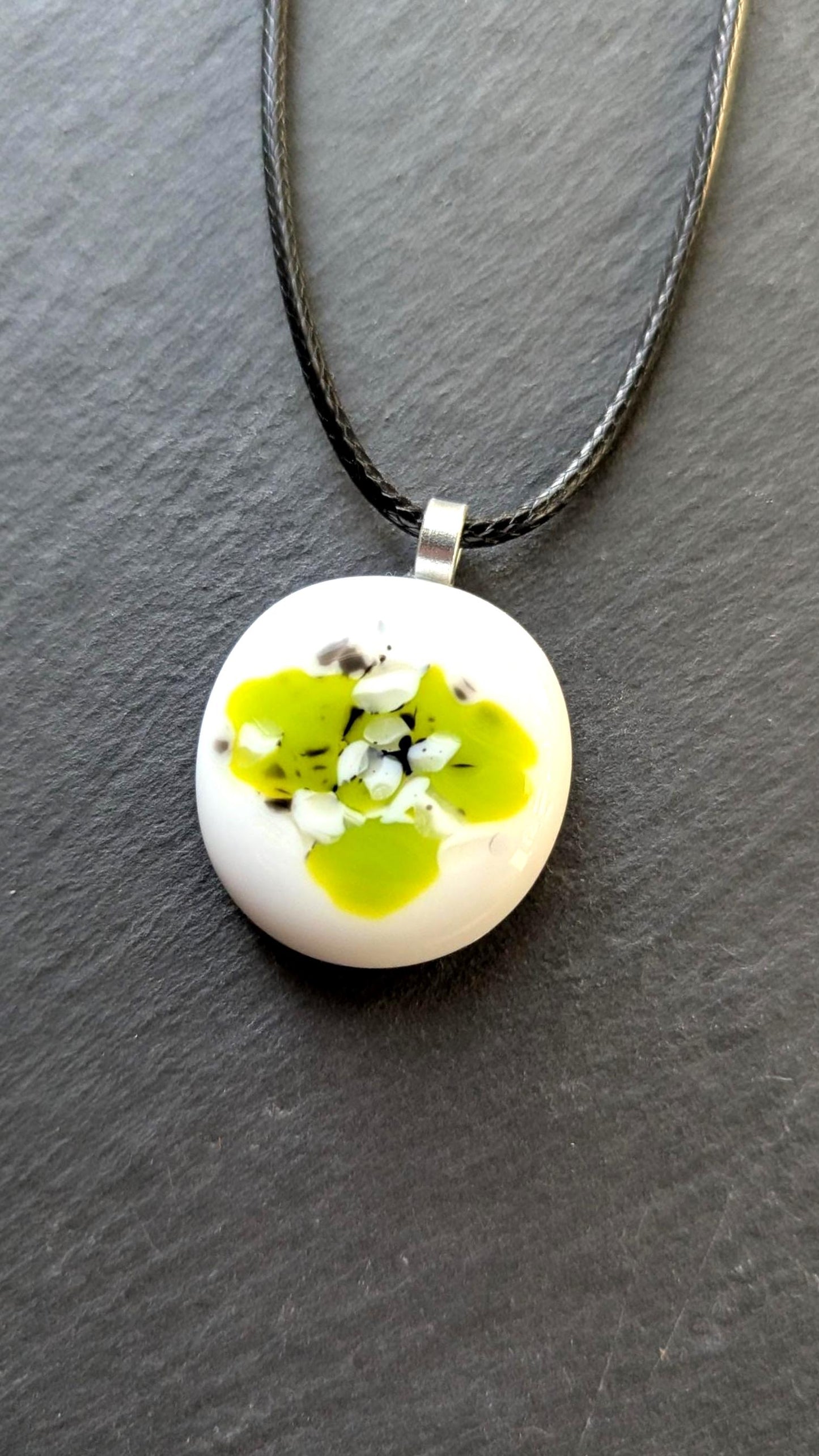 Round white fused-glass pendant with lime-green floral pattern on a silver bail, hanging from a black cord necklace.