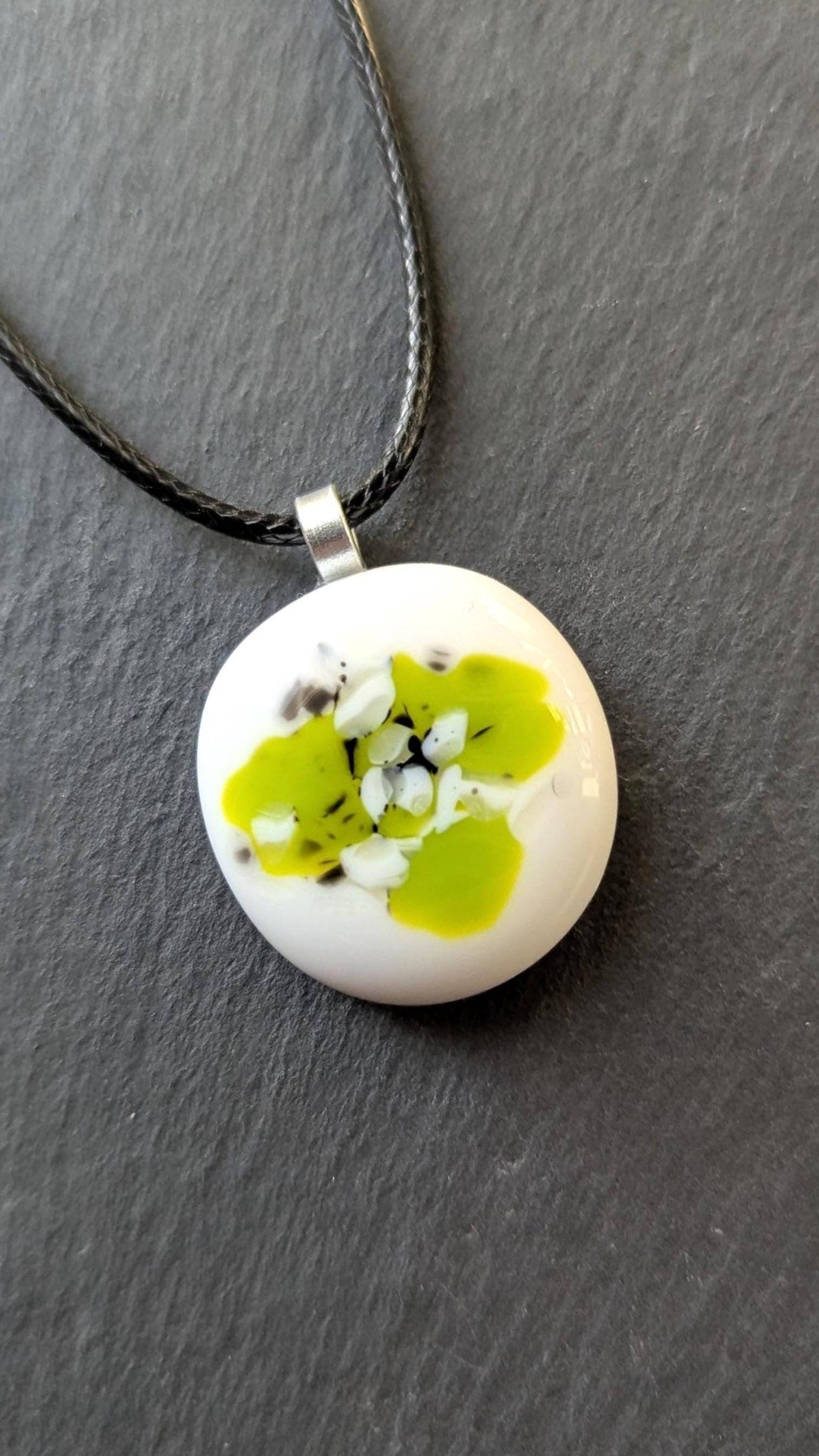 Round white fused-glass pendant with lime-green floral pattern on a silver bail, hanging from a black cord necklace.