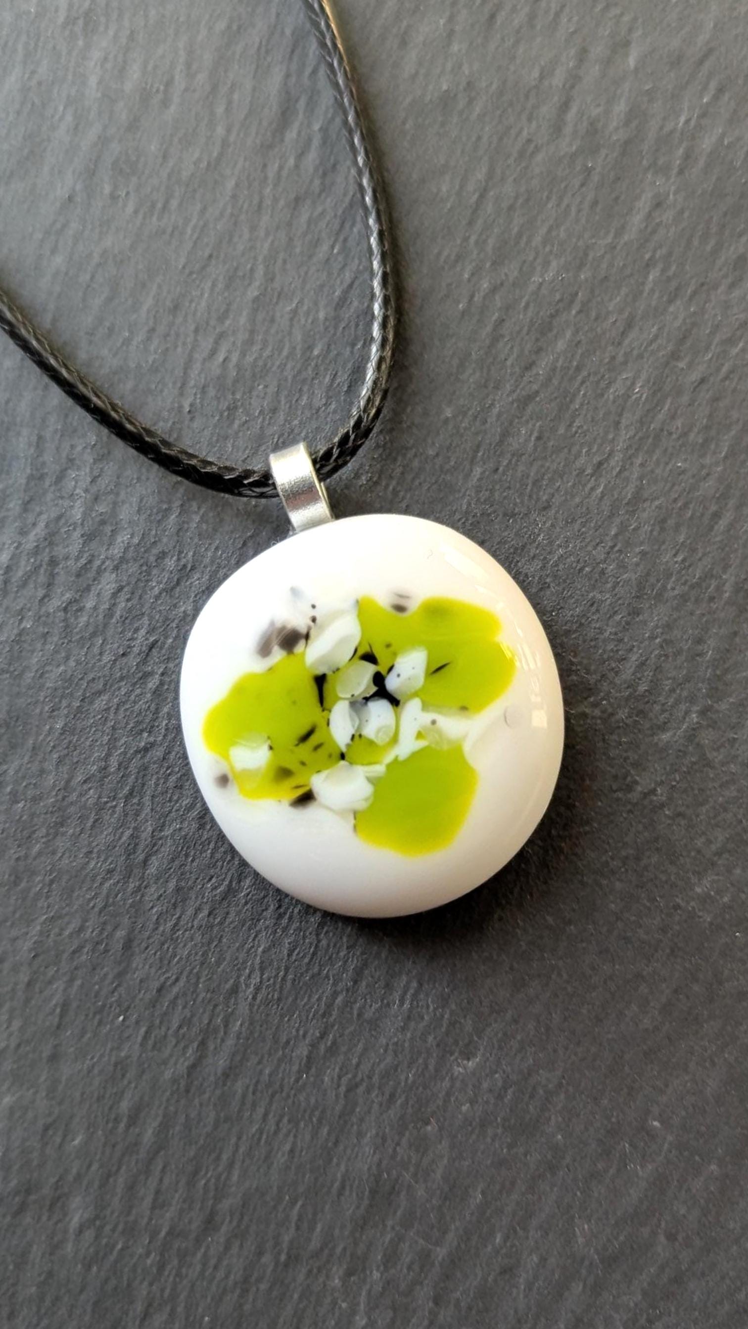 Round white fused-glass pendant with lime-green floral pattern on a silver bail, hanging from a black cord necklace.