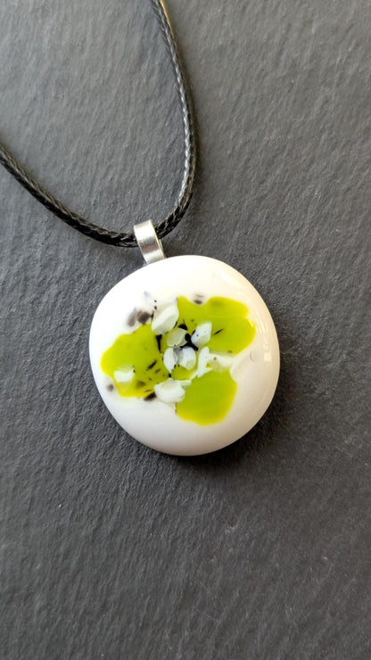 Round white fused-glass pendant with lime-green floral pattern on a silver bail, hanging from a black cord necklace.