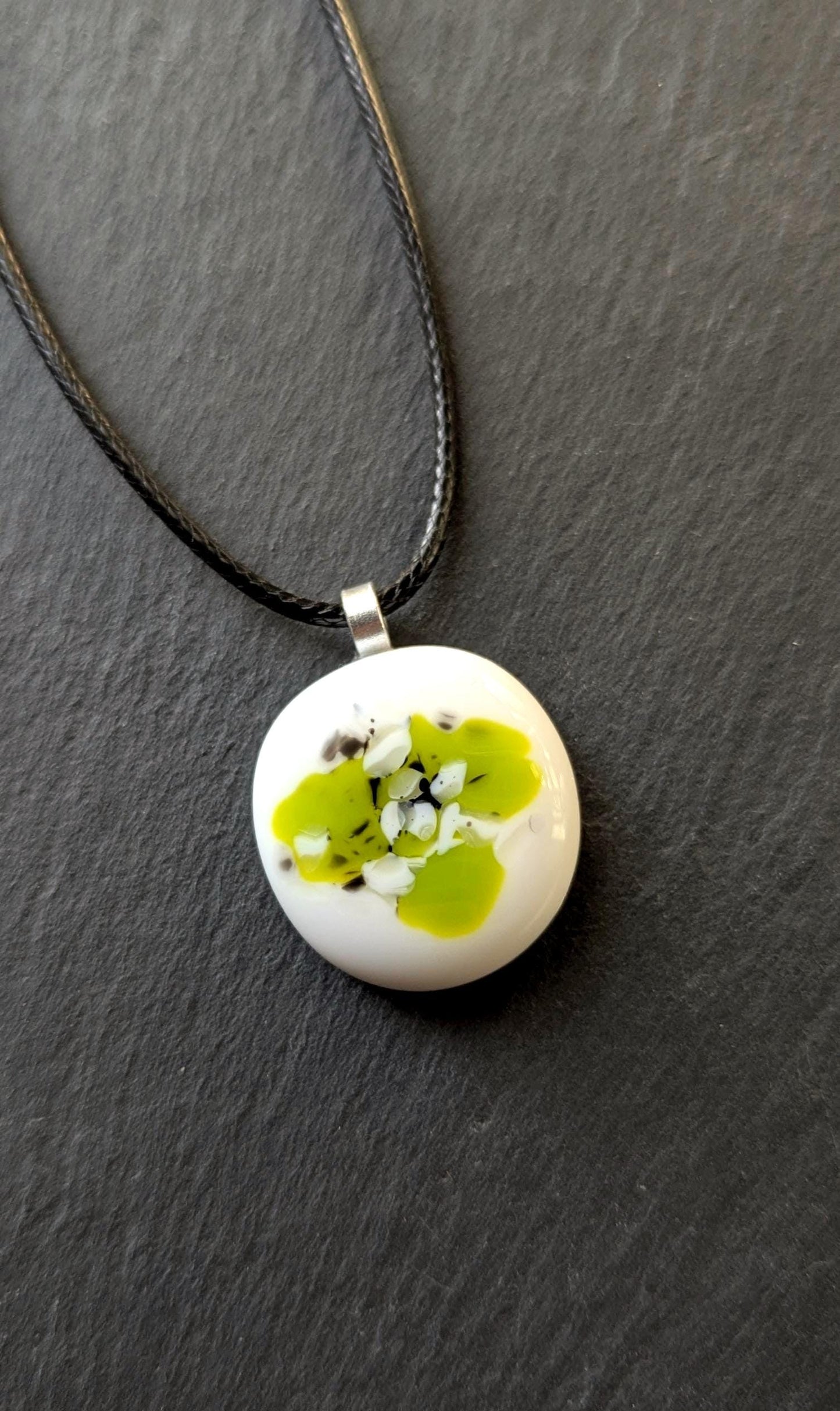 Round white fused-glass pendant with lime-green floral pattern on a silver bail, hanging from a black cord necklace.