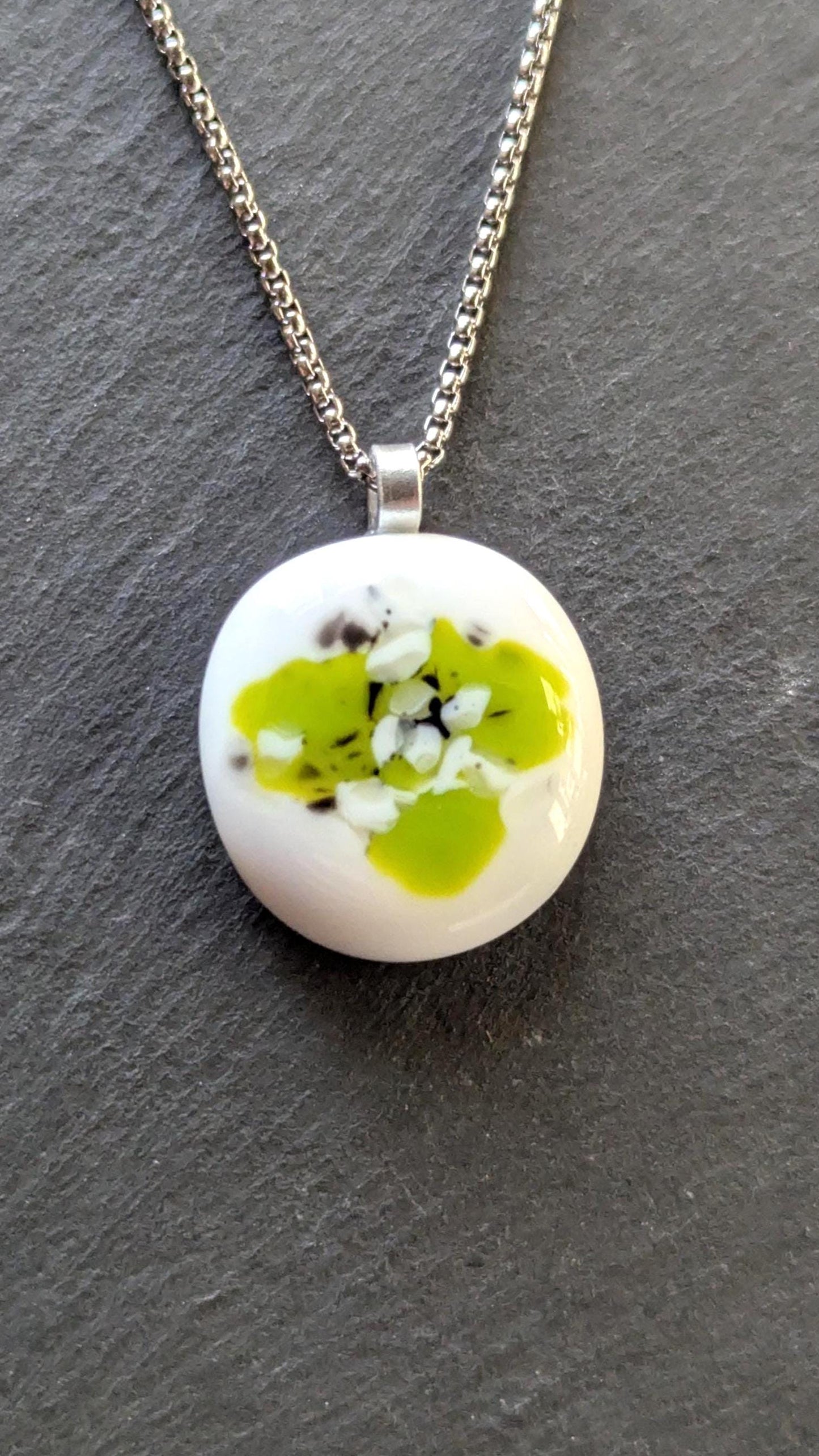 Round white fused-glass pendant with lime-green floral pattern on a silver bail, hanging from a black cord necklace.