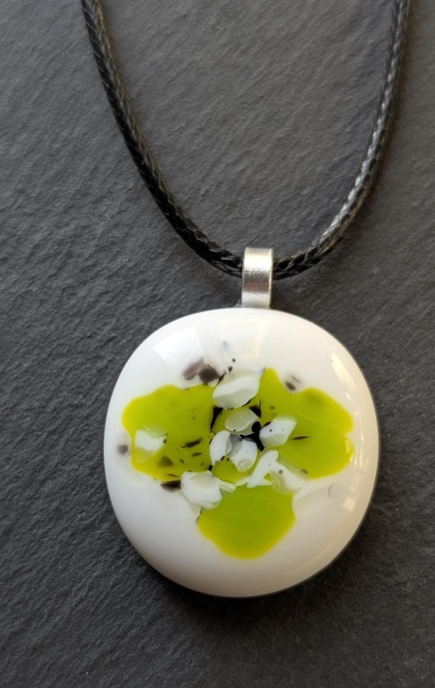 Round white fused-glass pendant with lime-green floral pattern on a silver bail, hanging from a black cord necklace.