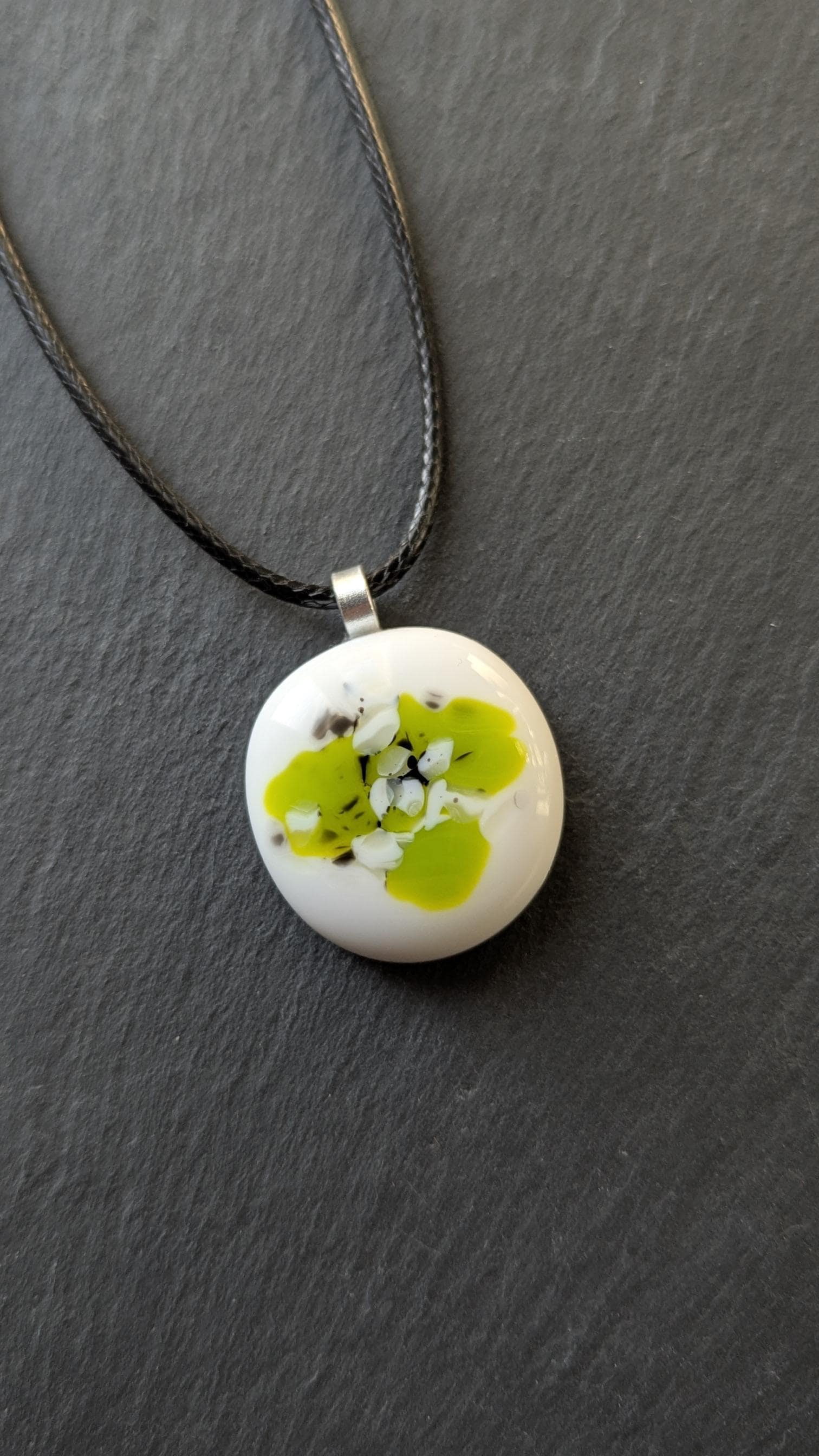 Round white fused-glass pendant with lime-green floral pattern on a silver bail, hanging from a black cord necklace.