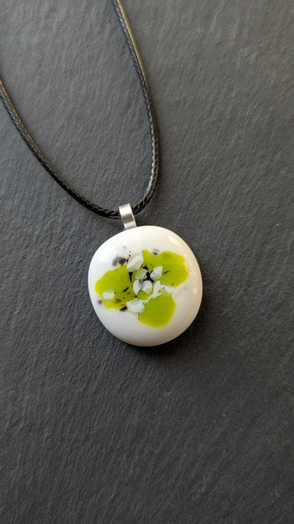 Round white fused-glass pendant with lime-green floral pattern on a silver bail, hanging from a black cord necklace.