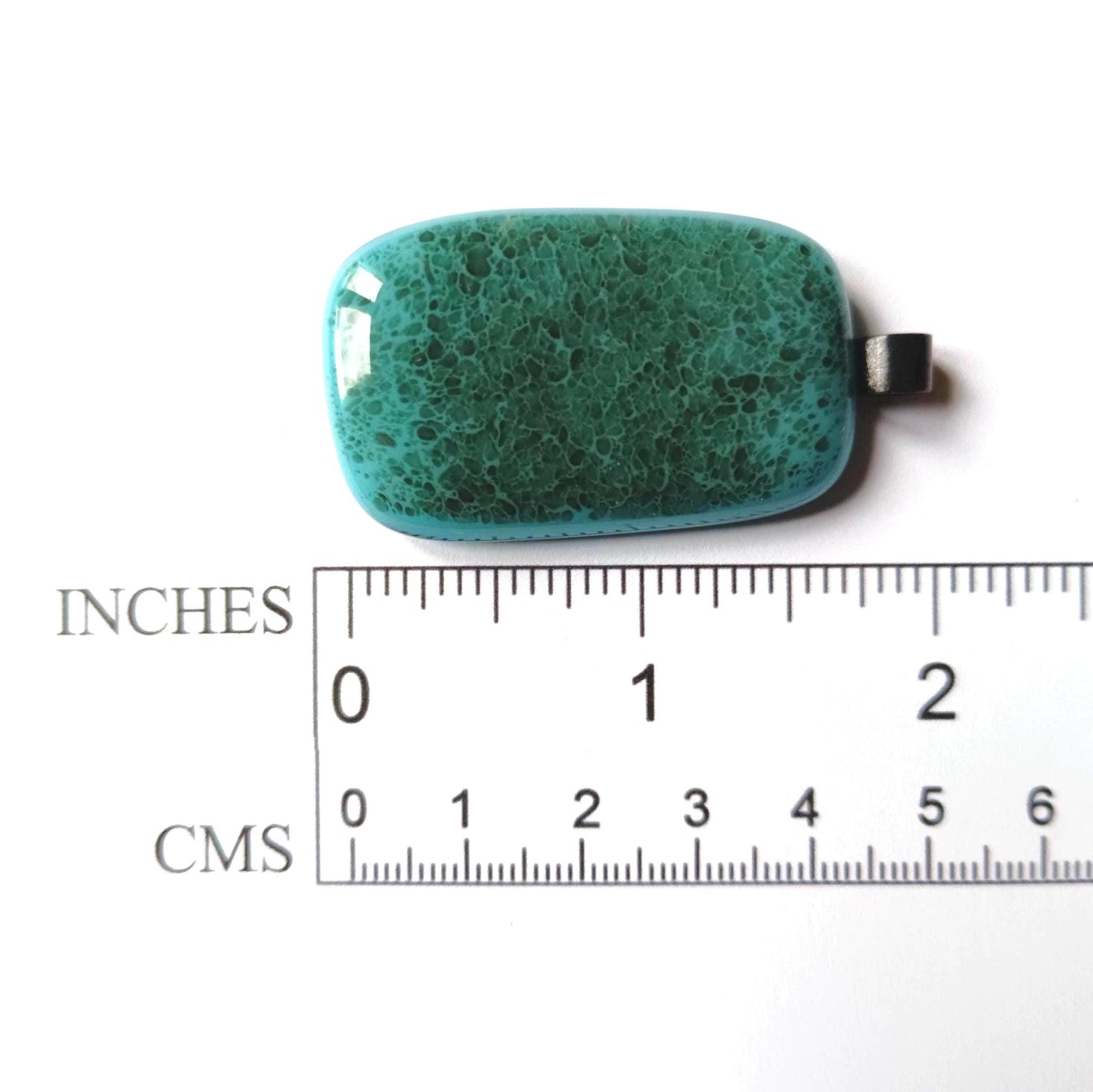A teal green glass pendant with a speckled texture. The pendant is approximately 1 inch long and 1/2 inch wide. The pendant is on a white surface with a ruler for scale.