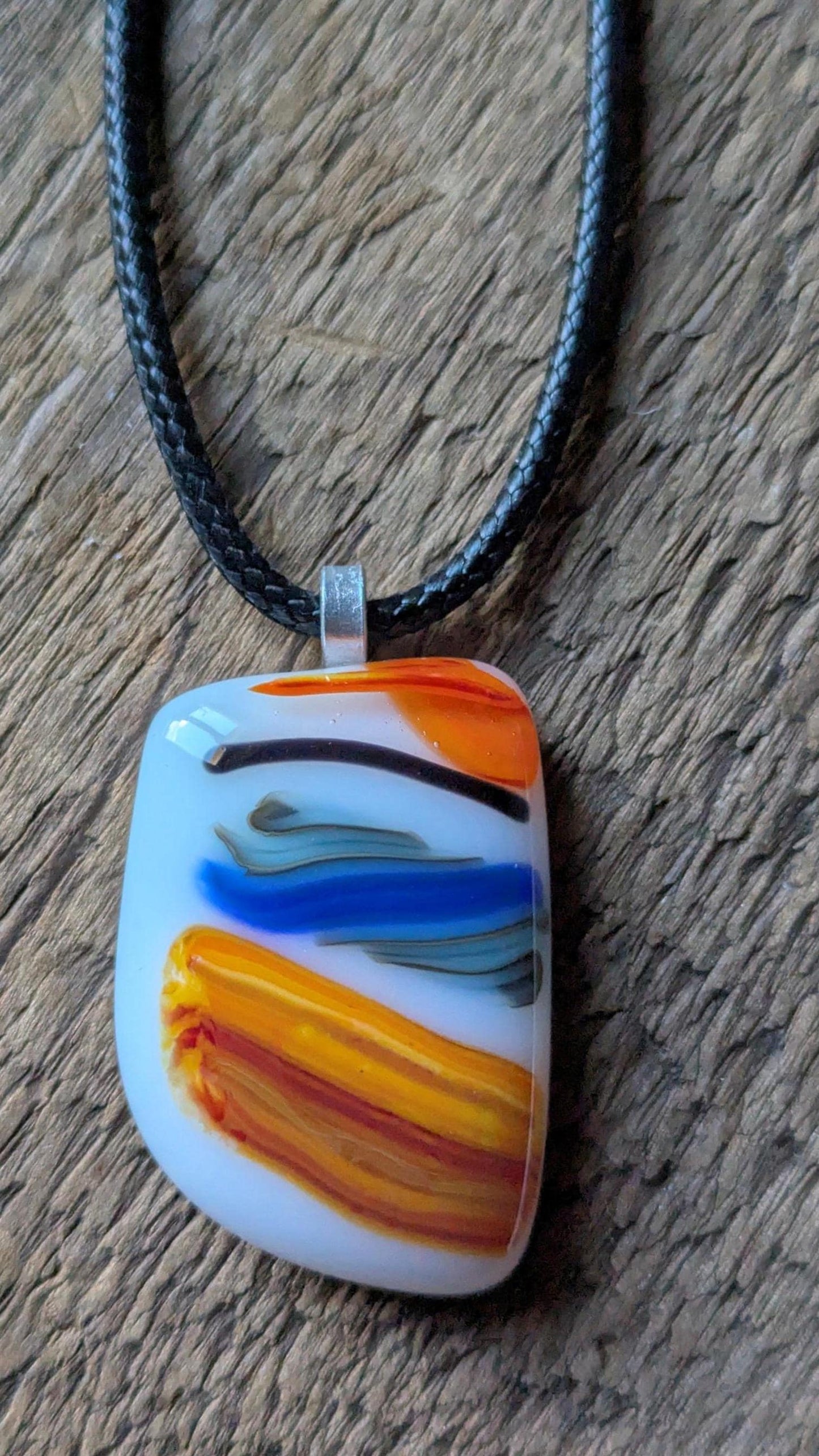 Large colorful fused glass pendant on black cord; white base with orange, yellow and blue swirls—handmade artisan jewellery.