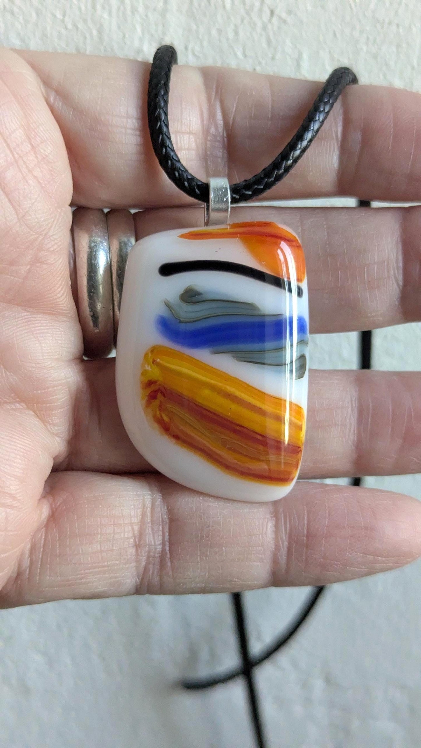 Large colorful fused glass pendant on black cord; white base with orange, yellow and blue swirls—handmade artisan jewellery.