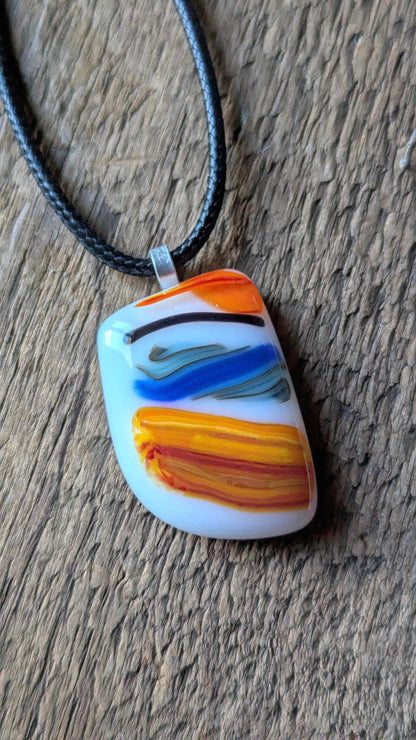 Large colorful fused glass pendant on black cord; white base with orange, yellow and blue swirls—handmade artisan jewellery.