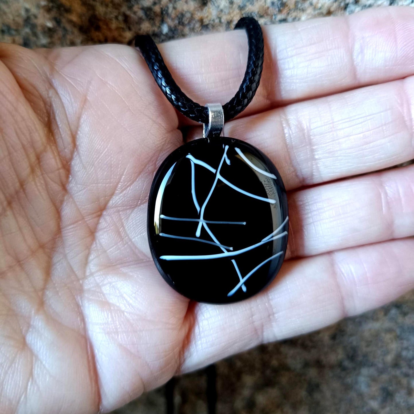 May include: A black glass pendant necklace with a white abstract design. The pendant is round and hangs from a black cord.