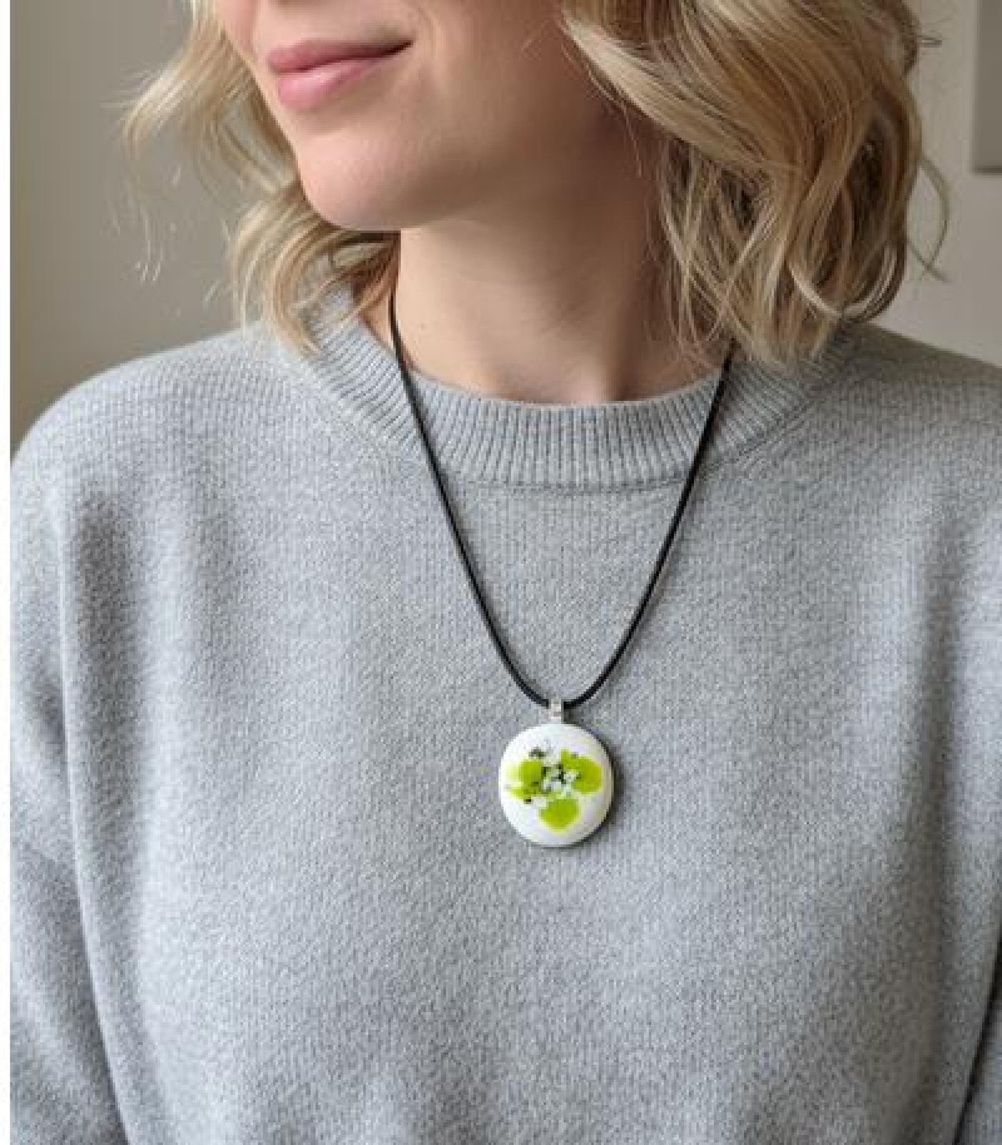 Round white fused-glass pendant with lime-green floral pattern on a silver bail, hanging from a black cord necklace.