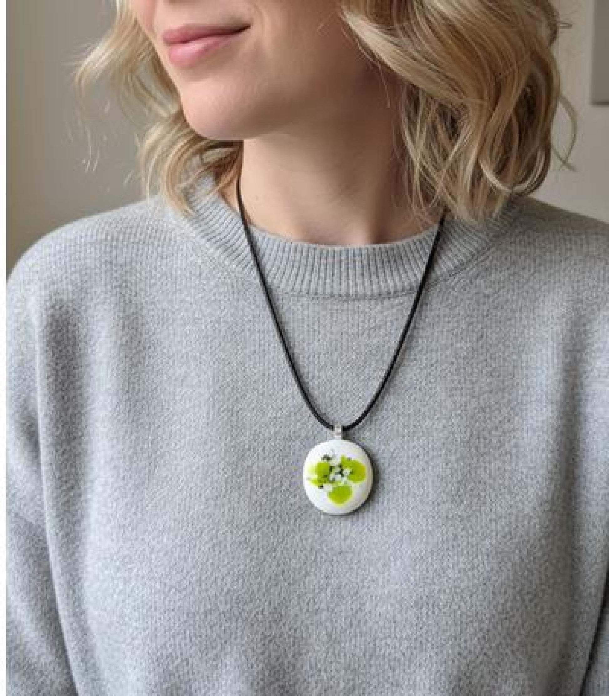 Round white fused-glass pendant with lime-green floral pattern on a silver bail, hanging from a black cord necklace.
