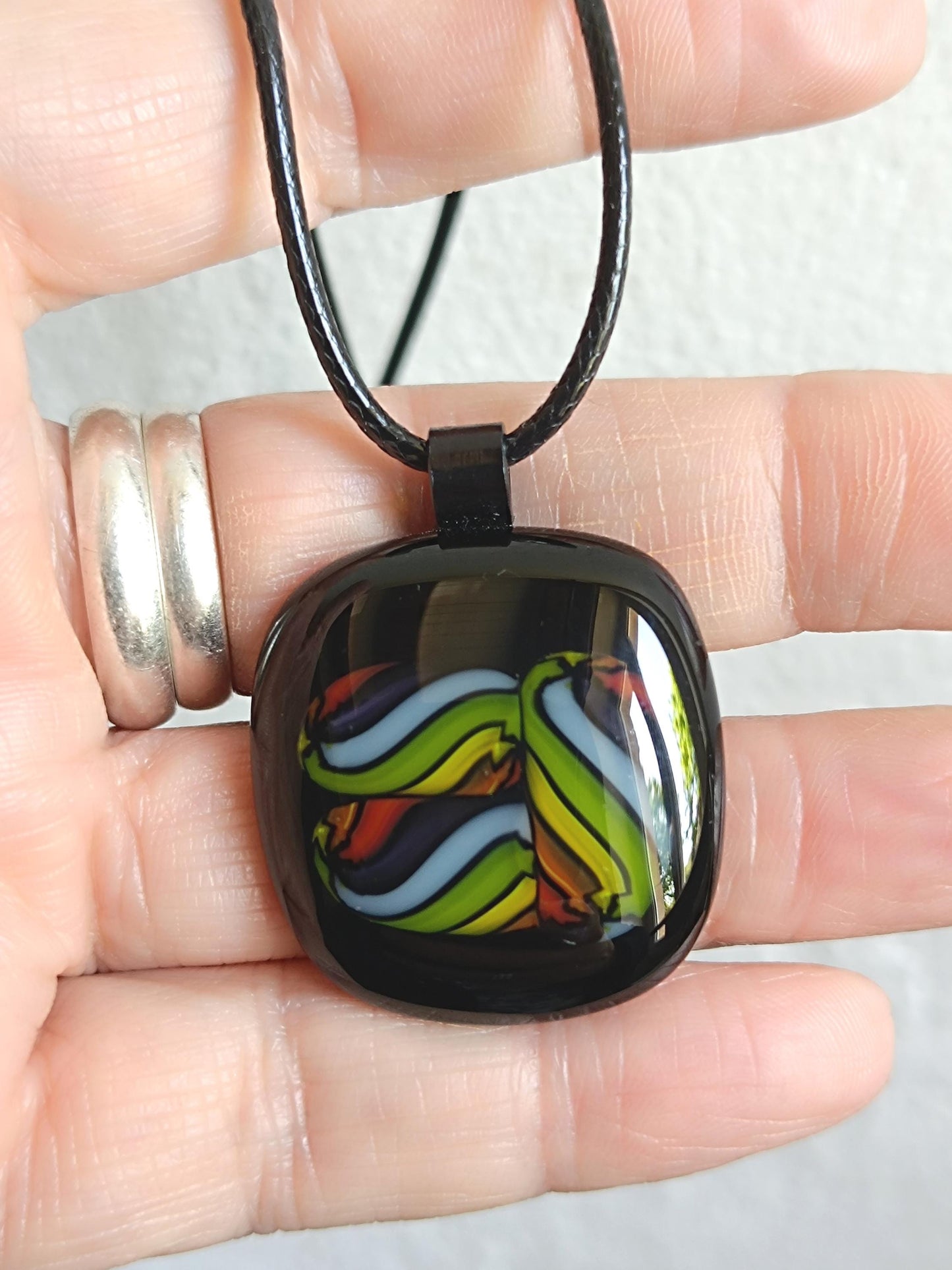 Square glass pendant in black bezel with multicolor swirl in green, blue, yellow, red on a black cord necklace.