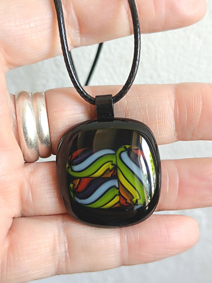Square glass pendant in black bezel with multicolor swirl in green, blue, yellow, red on a black cord necklace.