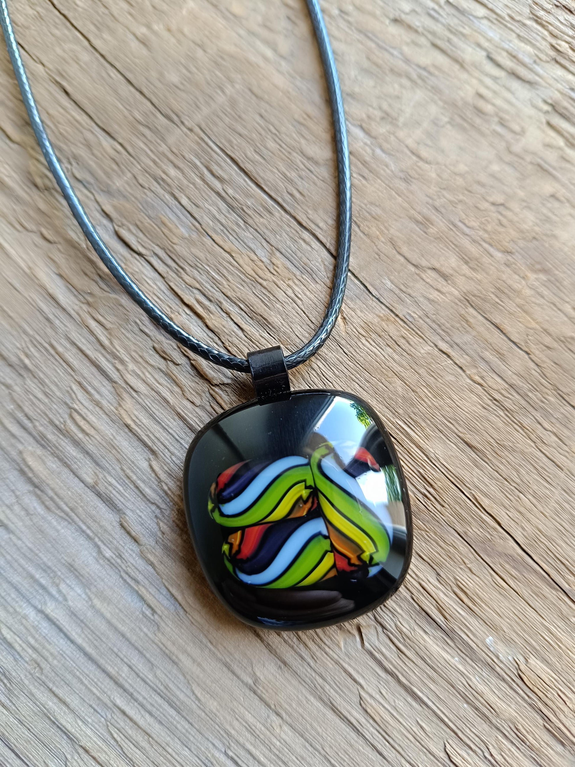 Square glass pendant in black bezel with multicolor swirl in green, blue, yellow, red on a black cord necklace.