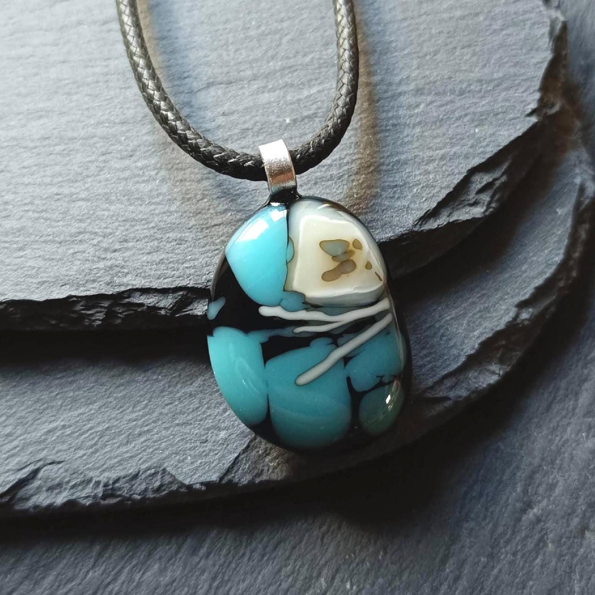 Fused glass pendant necklace in blue and black tones with white swirls and a silver bail on a black cord.