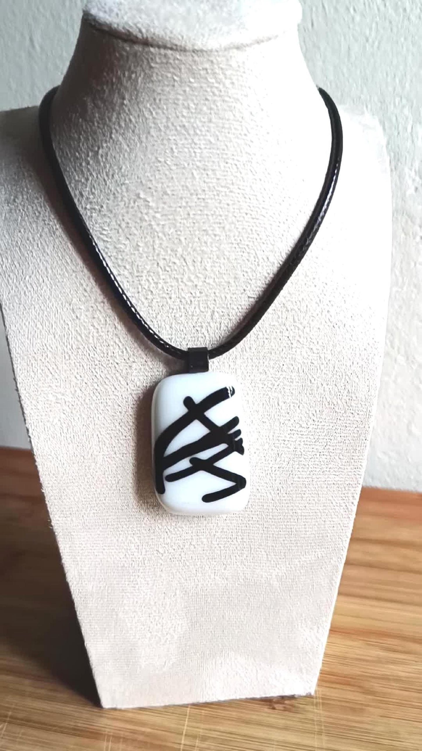 May include: A black cord necklace with a white glass pendant featuring black abstract lines.