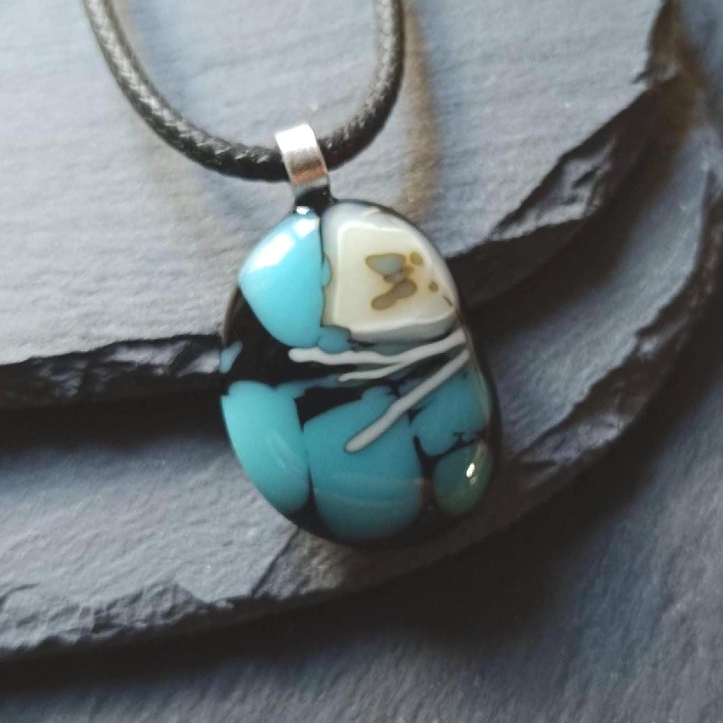 Fused glass pendant necklace in blue and black tones with white swirls and a silver bail on a black cord.